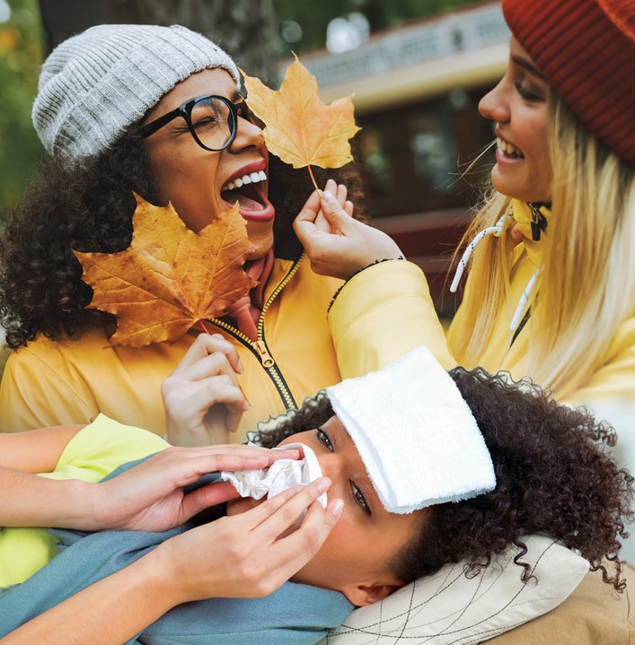 Two people dressed in autumnal clothes, holding orange leaf. Person at bottom of image is laid in bed with flu